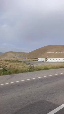 The video opens with a view of a desolate landscape featuring a paved road in the foreground and a barren, tan-colored hill in the background. White modular buildings are situated on the ground near the base of the hill. Large white letters spelling "НАРЫН РЕНОБАЦИЯ" are inscribed on the hillside. A truck with a rusty blue trailer drives by, partially obscuring the view of the buildings. The camera continues to pan, focusing on the hill with the inscription and the surrounding dry grass.