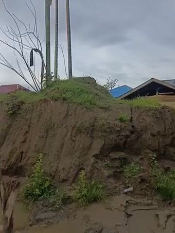 The video shows a landscape with evidence of a landslide or erosion. A steep, muddy embankment dominates the foreground, with visible signs of soil displacement. Several houses, some appearing damaged or under construction, are visible in the background, situated on higher ground or behind the embankment. Palm trees and other vegetation are scattered throughout the scene, some of which are partially obscured by the mud. The sky is overcast, suggesting possibly recent rainfall or a storm. The overall impression is one of environmental disturbance and potential damage to the structures.