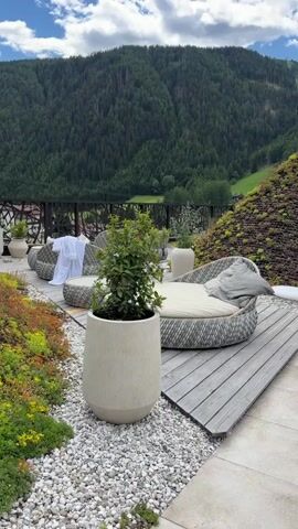 The video pans across an outdoor patio area with a scenic mountain view. In the foreground, a large, light-colored planter holds a green shrub. To the right, a round, cushioned lounger with gray upholstery sits on a wooden deck. In the background, a curved sofa with cushions and a white robe draped over it is visible, along with other planters and landscaping. A railing separates the patio from a densely forested mountain slope. The sky is partly cloudy.