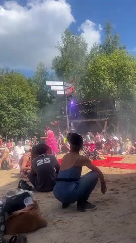 The video shows an outdoor event with a small stage set up in a clearing surrounded by trees. The ground is sandy. In the foreground, two people are sitting in a squatting position, facing away from the camera towards the stage. The person on the right is wearing a blue jumpsuit and a head covering. The person on the left is wearing a dark t-shirt with the number 25 on the back. A large brown bag and other items are on the ground near them. The stage area is elevated, with a wooden structure and a red carpet. A performer in a pink outfit is visible on the stage, interacting with an audience. Several people are seated and standing around the stage, watching the performance. Some are holding drinks or bags. The sky is blue with scattered clouds, and sunlight is filtering through the trees. There appears to be some mist or smoke on the stage.