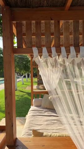 The video pans across a wooden cabana with white sheer curtains and a bed covered in a patterned throw. Outside the cabana, there is a swimming pool surrounded by green grass and trees. The sun is shining, casting shadows on the ground.
