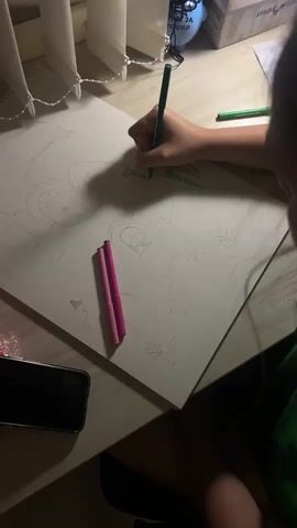 A close-up shot shows a person's hands as they draw on a large sheet of white paper. The drawing depicts architectural elements that appear to be buildings with arched doorways and windows, and some decorative floral or plant-like designs. The artist is using a green pencil to add detail to a grassy area in the drawing, with some pink markers also visible on the paper. A smartphone lies on the desk to the left of the drawing. In the background, a desk lamp casts a warm glow, and a window with vertical blinds is visible. The scene suggests a quiet, indoor activity, likely at a desk, possibly in a home or studio setting. There is some text on a blue object in the background that is partially obscured and difficult to read, but appears to be in Cyrillic script. The overall lighting suggests it is either early morning or evening.