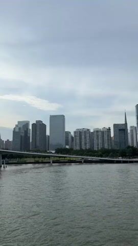 This media file showcases a panoramic view of a modern cityscape situated along a wide river. The scene captures a collection of towering skyscrapers, reflecting the architectural advancements of the location. Among the notable structures are the Canton Tower, a prominent landmark with its distinctive lattice design, and a modern stadium with a white, curved roof.

The river in the foreground appears calm, with its surface reflecting the overcast sky and the buildings on the opposite bank. A bridge spans the river in the middle ground, with visible traffic and pedestrian activity, suggesting a bustling urban environment. Along the riverbanks, trees and green spaces provide a contrast to the built environment, and a promenade with numerous people enjoying the view is visible in the latter part of the sequence.

The weather appears to be cloudy, with a soft, diffused light, indicating it might be late afternoon or early morning. The overall mood is one of urban vitality and modern progress. The location is identified as Yuexiu District, China, further contextualizing the impressive skyline. There is some visible text on the buildings, though it is largely illegible in the provided frames. The activity depicted is primarily scenic viewing and general city life.