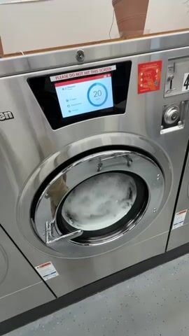 A close-up shot shows the interior of a commercial washing machine filled with white suds and laundry. The washing machine has a metallic exterior and a digital display screen showing the word "Normal" and a timer counting down from 20 minutes. The washing machine is in a laundry room with a tiled floor and a light-colored wall in the background.