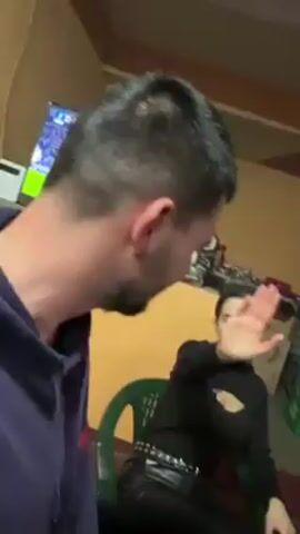 The video shows a group of people sitting around a table indoors, seemingly socializing. Initially, a woman in a black top and leather pants sits on a green chair, gesturing to stop the camera. The camera then pans to show three men at the table, drinking from glasses, and one man eating from a small bowl. There are bottles of alcohol, crates of drinks, and decorative pictures on the wall. The room appears casually decorated.