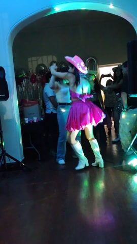 The scene is a lively party, probably a 20th birthday celebration, given the prominent balloon numbers in the background. The focus is a young woman in a vibrant pink dress and white boots dancing energetically under flashing lights. She also wears a fuzzy pink halo headpiece. The dance floor is dark wood. Other partygoers are seen in the background, some dancing and others watching. A table with party supplies and drinks is visible to the side. The atmosphere is festive and upbeat.