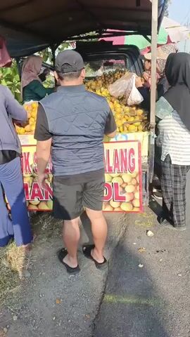 A group of people are gathered around a cart filled with oranges. A man wearing a vest and shorts stands with his back to the camera, looking at the oranges. Two women, one wearing a hijab and a green shirt, and another wearing a hijab and a patterned skirt, are also visible. The cart has a banner with Indonesian text and images of oranges. The ground is paved and appears to be a roadside. Sunlight suggests it is daytime.