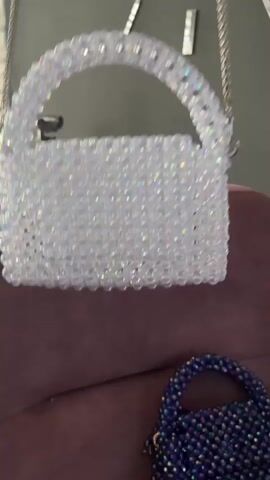 The video shows two beaded handbags against a backdrop of a couch and a wall with decorative elements. The larger handbag is white and clear, made of translucent beads, and is suspended by a silver chain. The smaller handbag, visible at the bottom right of the frame, is dark blue and also made of beads, with a portion of its handle showing. The scene is lit from the front, casting shadows and highlighting the reflective surfaces of the beads.