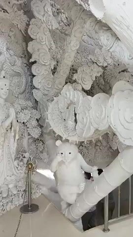 The video displays intricate white sculptures of figures and mythical creatures, possibly within a temple or artistic installation. In the foreground, a white bear-like sculpture is perched on a branch, reaching towards its mouth. To the left, two serene female figures, likely deities or important characters, stand side-by-side, adorned with delicate details and appearing to hold their hands in a gesture of reverence. Behind them and above, a large dragon sculpture winds its way through cloud-like formations, emphasizing the fantastical nature of the artwork. The overall environment is characterized by its monochromatic palette and the high level of detail in the sculpted elements. A metal stanchion with a golden ball sits in front of the female figures, suggesting a boundary or display marker. A glimpse of a person in gray clothing can be seen in the lower right corner, partially obscured by the sculptures. The lighting appears bright, highlighting the textures and forms of the white sculptures against what seems to be a darker background in some areas. The camera moves slightly, offering panning views of the artwork.
