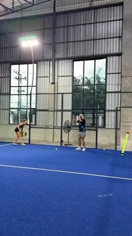 Two individuals are on a blue indoor padel court. The person on the left, a woman wearing athletic attire, is bent over, holding a padel racket. The person on the right, a man wearing a cap, shorts, and a t-shirt, is standing upright and appears to be instructing or demonstrating something to the woman. Several yellow padel balls are visible on the court. The court is enclosed by glass walls and metal fencing, with large windows in the background revealing an outdoor environment. Industrial-style lighting fixtures are mounted on the ceiling. A standing fan is positioned near the man.
