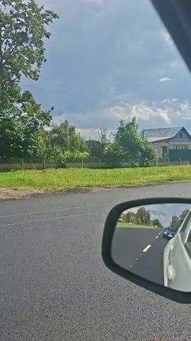 The video shows a view from inside a car, looking out the side window and at the side mirror. The road is asphalt, and there is a grassy area with trees and houses on the right side of the road. The sky is cloudy. The side mirror reflects the road behind the car, showing another car in the distance. The video appears to be taken while driving.