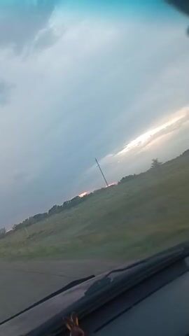 This video shows a view from inside a vehicle, looking out at a rural landscape. The camera is tilted and the footage is slightly blurred due to motion. The sky is overcast with some patches of sunlight breaking through. A long pole, possibly a utility pole or part of a wind turbine, is visible in the distance. The ground appears to be covered in grass and some scattered trees line the horizon. The scene suggests a drive through the countryside during a cloudy day with some sunlight.