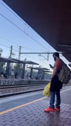 The video shows a train station platform with a man standing on the edge of the platform, looking at his phone. He is wearing jeans, an orange shirt, and orange shoes. He is also carrying a backpack and a yellow shopping bag. There are other people sitting and standing on the opposite platform, waiting for the train. There is graffiti visible on the wall behind the tracks. The sky is light blue.