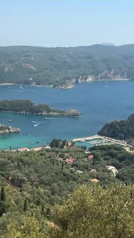 The video presents a sweeping aerial view of a picturesque coastal landscape, likely a bay or harbor, under a clear, bright sky. The scene is dominated by the deep blue of the sea meeting lush green, tree-covered hills that form a dramatic backdrop. In the foreground, foliage partially obscures the view, adding a sense of depth.

Closer to the water, a small village or settlement is visible, characterized by terracotta-tiled roofs nestled amongst the trees. A marina or harbor area is clearly defined, with numerous boats moored in the calm, turquoise waters. Some of these boats appear to be small yachts or fishing vessels. There are also a few structures along the waterfront, possibly restaurants or shops.

Further out in the bay, several smaller boats are actively moving, leaving white wakes on the water's surface, suggesting recreational activities like water skiing or leisurely cruising. The coastline is irregular, with rocky outcrops and small coves visible along the edges of the larger bay.

In the distance, the sea stretches out to the horizon, meeting a pale blue sky. The overall impression is one of a serene and idyllic Mediterranean or similarly warm climate location, likely a popular tourist destination given the presence of the marina and evident activity on the water. There are no people directly visible in the frame, but the evidence of human presence is clear through the buildings, boats, and infrastructure. The terrain is hilly, with winding roads visible in some areas, indicating accessibility to the settlement.