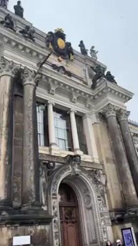 The video shows the exterior of a stately building. The building has an arched entrance with large wooden doors, flanked by Corinthian columns and ornate sculptures. The facade is detailed with numerous windows, decorative moldings, and statues atop the building. There is a golden coat of arms above the entrance. The sky is overcast and snow is visible on the ground. A few people are walking in front of the building.  In the background, the spires of other buildings are visible, suggesting the location is a historic city.