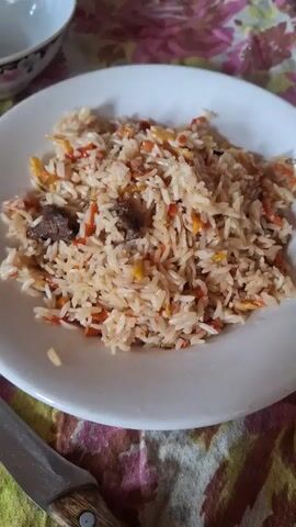 The video shows a white plate filled with rice pilaf, likely an Uzbek version, featuring rice, meat chunks, shredded carrots, and some yellow elements. The plate rests on a colorful floral patterned tablecloth next to a knife. A small white bowl is visible in the upper left corner. The camera zooms in slightly on the pilaf.