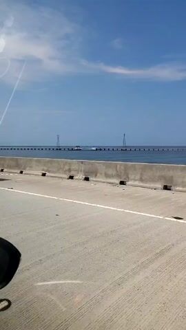 The video shows a view from a vehicle driving on a road with a concrete barrier. In the background, there is a long bridge over water, supported by multiple pillars. Along the bridge are tall, metal electrical towers. The water is calm and blue under a partly cloudy sky. The perspective suggests the viewer is looking out the side window of the vehicle.