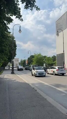 This is a daytime video, shot from a sidewalk, looking down a city street in Munich, Germany. The sky is partly cloudy, suggesting fair weather. Trees line the left side of the street, casting shadows on the sidewalk. On the right, a modern, white building with vertical lines dominates the scene. A street lamp with an ornate design stands on the left.

The street is populated with vehicles, indicating normal daytime traffic. Cars of various models, including a white Smart car, a silver SUV, and a white station wagon, are visible on the road. A white van is also present. There are no people clearly visible in the foreground or immediately engaged in activity. The overall mood is neutral, depicting an everyday urban environment. The street appears to be a public thoroughfare with buildings and trees suggesting a city setting, possibly a commercial or residential area. The presence of a rainbow flag visible in the distance adds a detail of potential cultural significance. No legible text is present on the vehicles or buildings.
