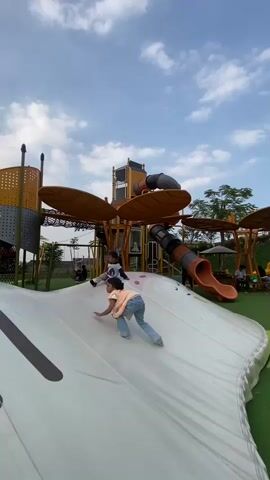 A low-angle shot captures a playful scene at an outdoor playground under a partly cloudy sky. In the foreground, a white, inflatable ramp dominates the frame, with a young girl in a light orange vest and blue jeans crawling up its slope. She is positioned in the lower center of the image, facing away from the camera. Further up the ramp, another child, mostly obscured, is also visible. The playground structure behind them is a complex, multi-level design featuring yellow and gray components, netting, and orange tube slides. The overall atmosphere is one of youthful activity and outdoor recreation.