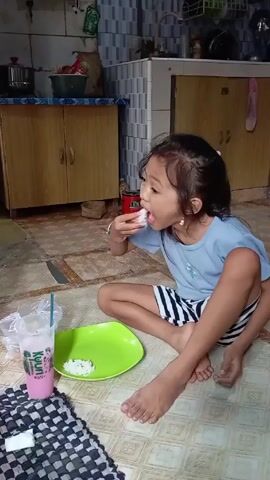 A young, barefooted girl sits cross-legged on a tiled floor in a kitchen setting, eating rice with her fingers from a bright green plate. She is wearing a light blue t-shirt and black and white striped shorts, and has a small ponytail. To her left is a cup of pink liquid with a straw and a black and white checkered mat. The kitchen features wooden cabinets, tiled walls, and various kitchen items in the background. The girl looks at the camera and makes a thumbs-up gesture.