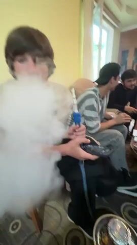 A group of five young men are gathered around a small, round table in a living room. The table is littered with green bottles of juice or beer, a few cans, cell phones, and a pack of cigarettes. One man in the foreground is holding a hookah pipe and exhaling a large cloud of smoke. The others are sitting in chairs or on a sofa, some looking at their phones and others seemingly engaged in conversation. The room is furnished with a wooden cabinet built into the wall, and there's a window letting in natural light. The walls are painted in a warm yellow hue.