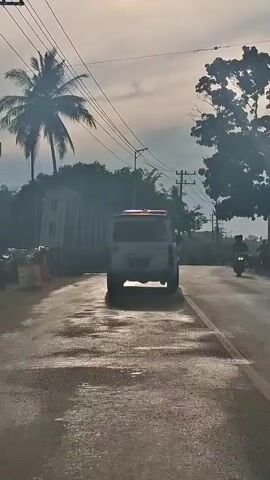 A white van, possibly an ambulance, is driving down a paved road. The road appears wet, with several puddles reflecting the light. To the left of the road, there is a wall and lush green vegetation, including palm trees. Overhead, telephone poles and power lines stretch across the sky, which is partly cloudy. On the right side of the road, there are trees and some buildings in the distance. A person on a motorcycle is also visible on the road. The sun is shining, casting long shadows and creating a bright, hazy atmosphere. The video is taken from a moving vehicle, likely another car, following the white van.