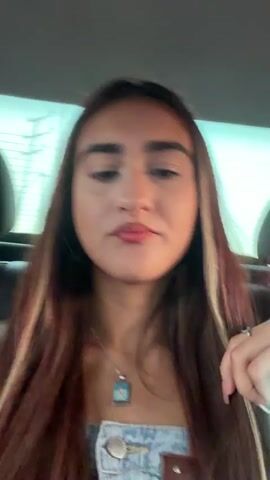 A young woman with long, brown hair, featuring blonde highlights, is filmed from the chest up. She is wearing a denim overall-style top with a silver necklace that has a light blue pendant. The background appears to be the interior of a car, with dark seats and a slightly blurred view of the outdoors through the windows. The woman pouts her lips and moves her head slightly, occasionally touching her hair or necklace. The lighting suggests it is daytime.