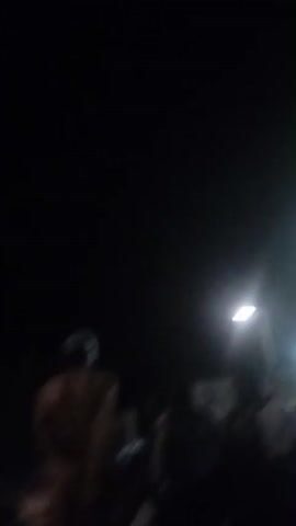 The video depicts a night scene in Zongoma Kyari, Nigeria, illuminated by a bright street lamp. Silhouetted figures of people and what appear to be motorcycles are visible in the foreground, suggesting a gathering or a busy street at night. Several other streetlights are also visible in the background, creating streaks of light due to the motion blur. The overall atmosphere is dark, with the strong artificial light from the lamp creating a halo effect. The blurriness of the footage, likely due to camera movement or low light conditions, obscures fine details of the people and objects, rendering them as indistinct shapes. There are no discernible weather conditions, but the darkness implies it is nighttime. No explicit text is legible in the video. The scene appears to be outdoors, possibly a public space like a market street or a gathering point. The activity seems to be people milling about or waiting, with the presence of motorcycles suggesting transportation or a local meeting point.