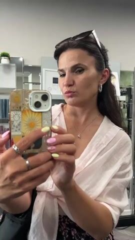 A woman with long dark hair, wearing sunglasses on her head, a light pink collared shirt tied at the waist, and a floral skirt, takes a selfie in a mirror. Her nails are painted pink and yellow. She is wearing rings on her fingers and earrings. She is holding a phone with a decorative case. In the background, there are shelves with various bottles and products, and mirrors. The woman makes a peace sign with her fingers.