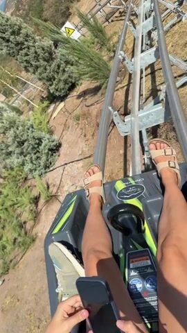 A first-person perspective video shows a person riding a mountain coaster. The coaster is on a track that winds through a dry, arid landscape with sparse vegetation. The person's legs and feet, wearing sandals, are visible in the foreground. The coaster is moving at a moderate speed, and the scenery changes as it navigates the track. In some shots, buildings and a lake are visible in the distance. The person occasionally reacts with sounds of excitement or surprise.