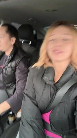 The video shows three people in a car, with the driver and front passenger visible, and a girl in the back. The driver is wearing a black vest over a purple top, and grey leggings, looking focused on the road. The front passenger is wearing a black jacket with a visible pink shirt underneath and smiles at the camera. The girl in the back is wearing a silver jacket and smiles. The car is moving, and loud music is playing.