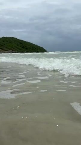 The video shows a low-angle view of waves gently lapping onto a sandy beach. The water is light green and frothy. In the background, a green, tree-covered hill slopes down towards the sea. The sky is overcast and cloudy. There are no people or other objects visible. The camera appears to be stationary.