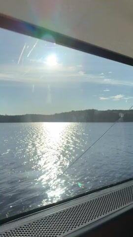 This video shows a view from a window, likely on a boat or vehicle, looking out at a body of water. The sun is shining brightly, creating a shimmering reflection on the water's surface. A tree-lined shore is visible in the distance. The bottom of the frame shows a portion of the interior, including a speaker grille and what appears to be a window frame.