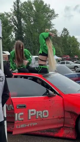 Two women in matching green tops and black pants sit on the roof of a red car, posing and interacting, in a car park at an event. The car, which has mud splattered on its lower panels, has "IC PRO ЦЕНТР" written on the door. A man in a grey jacket stands by the car, while other people mill about in the background. There are trees visible beyond the parked cars, and the sky is overcast.