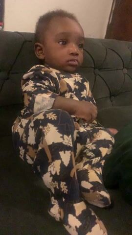 A baby wearing a patterned sleeper sits on a dark green buttoned couch, looking attentively toward the left. The baby has dark skin and short, dark hair. The sleeper is cream, tan, and dark blue, with an autumnal leaf pattern. The couch appears to be in a living room.