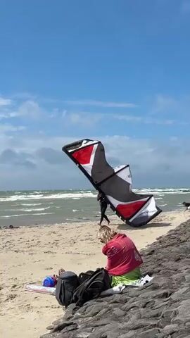 A kitesurfer wearing a black wetsuit and helmet stands on a sandy beach near the ocean. The kitesurfer is holding a large, multi-colored kite, which is folded and partially deflated. The kite is primarily black, white, and red. The ocean is visible in the background with choppy waves and a cloudy sky. In the foreground, a person with blonde hair wearing a pink and red towel is sitting on the sand, facing away from the camera. Other beach accessories, including bags and a blue object, are also visible in the foreground. The kitesurfer appears to be preparing to launch or disassembling their equipment.