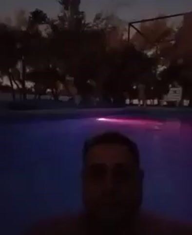 A man takes a selfie video while standing in a swimming pool at dusk. The pool is lit with a red light. The surrounding area includes trees, a basketball hoop, lounge chairs, and a camper trailer. The sky is visible in the background, suggesting it is either late evening or early morning. The man is looking at the camera with a neutral expression. The video appears to be stable.