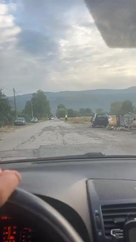 The video, shot from inside a vehicle, shows a road with visible potholes. A figure dressed in a high-visibility vest and dark pants stands by the side of the road. The figure is positioned next to a tall, dark rectangular object with yellow markings and a blue bucket at its base. In the background, there are trees, fields, and distant mountains under a cloudy sky. Several cars are visible on the road and parked to the side. The driver's hand is visible on the steering wheel.