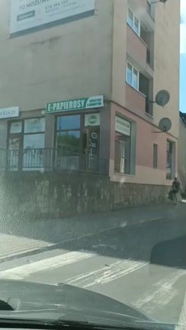 A person walks down a street, carrying several bags. The street is lined with buildings, some of which have shops on the ground floor. The sky is blue with white clouds. The video appears to have been taken from inside a car.