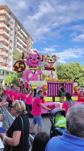 A brightly colored parade float, adorned with flowers, depicts two cartoon dogs. One dog, pink, has a large bone with a circular design on it extending from its back. The other dog, tan, is seated next to the pink dog. The float also features a pink and yellow structure with bars, and on it, a young girl in a green outfit is dancing. People in bright pink shirts and green scarves walk alongside the float, some interacting with the crowd. Spectators, including a woman with blonde hair and a man in a blue shirt, are visible in the foreground. The scene takes place outdoors on a sunny day, with a tall apartment building in the background.