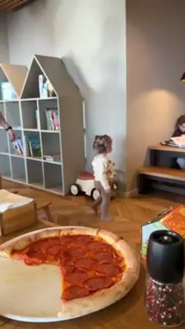 The video shows a man standing in a restaurant or cafe. On the table in front of him is a large pepperoni pizza with a few slices missing. A toddler with pigtails is walking around the room. Behind the man, there are house-shaped bookshelves and a small toy car. There is also a table with a bench where a young girl is sitting and looking at a book. The man appears to be watching the toddler, perhaps concerned about her safety.