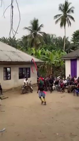 The scene captures an open area with a performer at its center, engaged in an energetic dance. The performer is adorned in a colorful, possibly ceremonial costume. A crowd encircles the dancer, seated in chairs and standing, observing the performance. The backdrop features two simple houses with corrugated roofs. Palm trees and lush greenery provide a verdant backdrop, suggesting a rural or tropical setting. Two motorcycles are parked near one of the houses. The overall setting indicates a cultural event or performance taking place in a village environment.