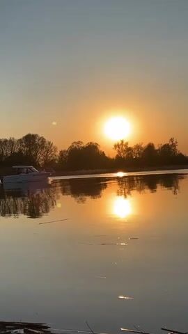 The video shows a small motorboat sailing on a calm lake at sunset. The sun is a bright circle in the sky, creating an orange and yellow glow. The sky is a pale blue. The water reflects the sun and sky, creating a mirror image of the scene. The boat is moving from left to right in the frame, and there are trees silhouetted in the background. A few pieces of driftwood are in the foreground.