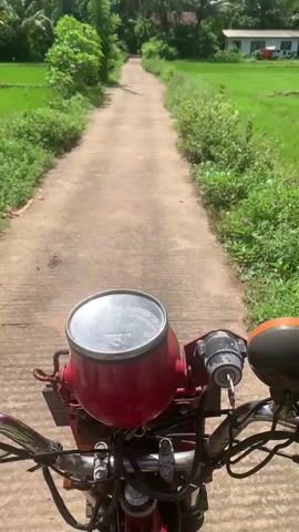 The video captures a motorcycle journey through a serene rural landscape in Karandugoda, Sri Lanka. Filmed from the perspective of the rider, the initial shot at 0:00 shows the motorcycle's red speedometer, marked from 0 to 80, with an odometer reading "99471" km and Japanese text visible below it. The motorcycle is on a narrow, unpaved road, flanked by vibrant green rice paddies on both sides. Lush tropical vegetation, including palm trees and dense bushes, borders the fields under a bright blue sky dotted with white clouds. As the video progresses (0:01-0:04), the camera pans across the expansive rice fields and the surrounding verdant foliage, highlighting the peaceful and verdant nature of the countryside.