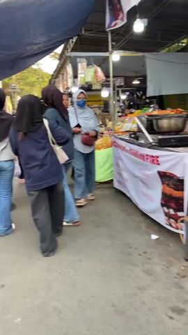 The video shows a street scene with several people walking by market stalls. In the foreground, a person in a blue headscarf and mask is walking towards the right. Behind them, to the left, a person in a black headscarf and blue shirt is walking away from the camera, with another person in a black headscarf beside them. Further back, a young man in a black t-shirt and shorts is walking, and to his right, a man in a pink shirt is walking. Market stalls with various goods are visible to the right and in the background. The overall environment appears to be an outdoor market.