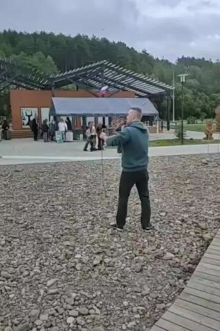 A man in a teal hoodie and black pants stands on a gravelly area, holding a rope with a small Russian flag attached to one end. He is swinging the rope in a circular motion, preparing to throw it. Behind him, a building with an open-air roof structure is visible, with several people gathered around tents and displays. The background consists of a forested hillside under a cloudy sky. A wooden boardwalk runs along the right side of the frame.