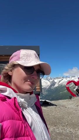 A woman is taking a selfie outdoors on a sunny day. She is wearing a pink baseball cap, sunglasses, and a pink puffer vest over a white jacket. Behind her, there is a modern-looking building with wooden accents, and beyond that, majestic snow-capped mountains are visible under a clear blue sky. To the right of the building, a large red heart-shaped sculpture with the word "DOMBAY" is visible. The ground is rocky and uneven.