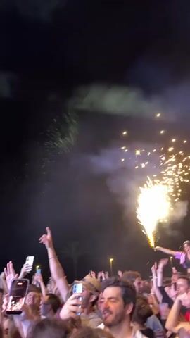 A crowd of people is gathered at an outdoor event, likely a concert, at night. Most of them are looking up and holding up their phones to record. Fireworks are going off in the background. As the camera pans up, three people are seen standing on each other's shoulders. The base of the structure is a man in a white shirt. On his shoulders is a woman in a purple crop top and jeans. On her shoulders is another woman wearing a striped top and black shorts. They are positioned near a street sign. The overall atmosphere is festive and celebratory.