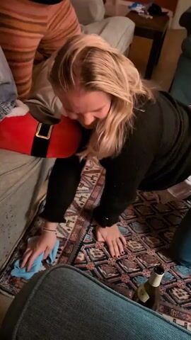 A woman with long blonde hair is on her hands and knees, using a light blue cloth to clean a patterned rug. She is wearing a black long-sleeved shirt. In the background, there are people sitting on a couch; one person is wearing orange and pink striped pants and another is wearing jeans with white socks. A red and black Santa-themed pillow is on the couch. A brown beer bottle is on the floor near the rug.  A dark brown table with various objects is in the background.