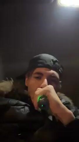 A young man in a black puffer jacket with a fur-trimmed hood and a backwards black baseball cap is in a car. He's holding a vaping device with a green light. He says something about being gassed, then waves his hand at the camera. He then smiles and continues to talk. The interior of the car is dimly lit.